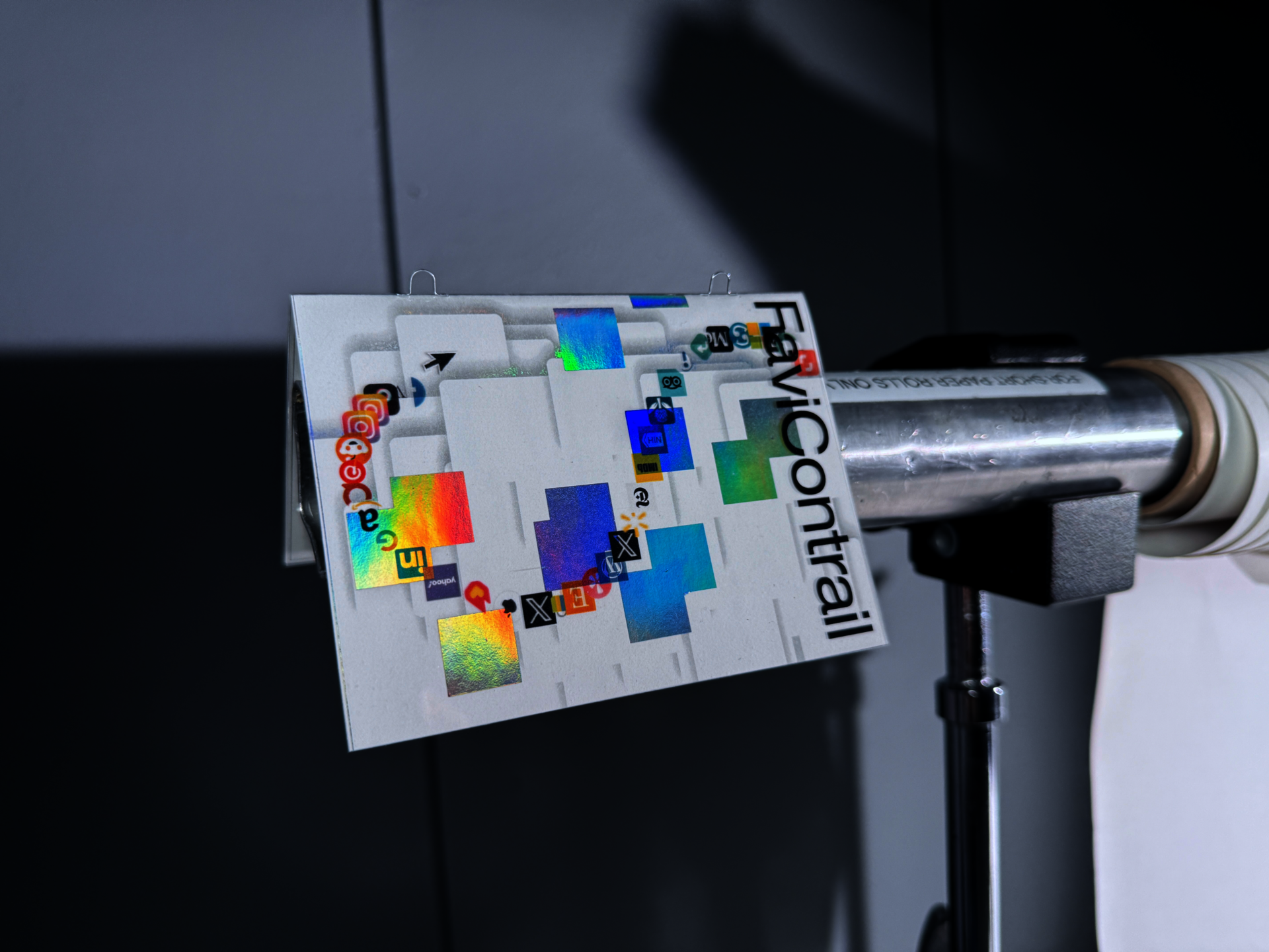 Close-up of the FaviContrail brochure print clipped to a metal pole, showing browser favicons trailing across layered white shapes and holographic color patches.