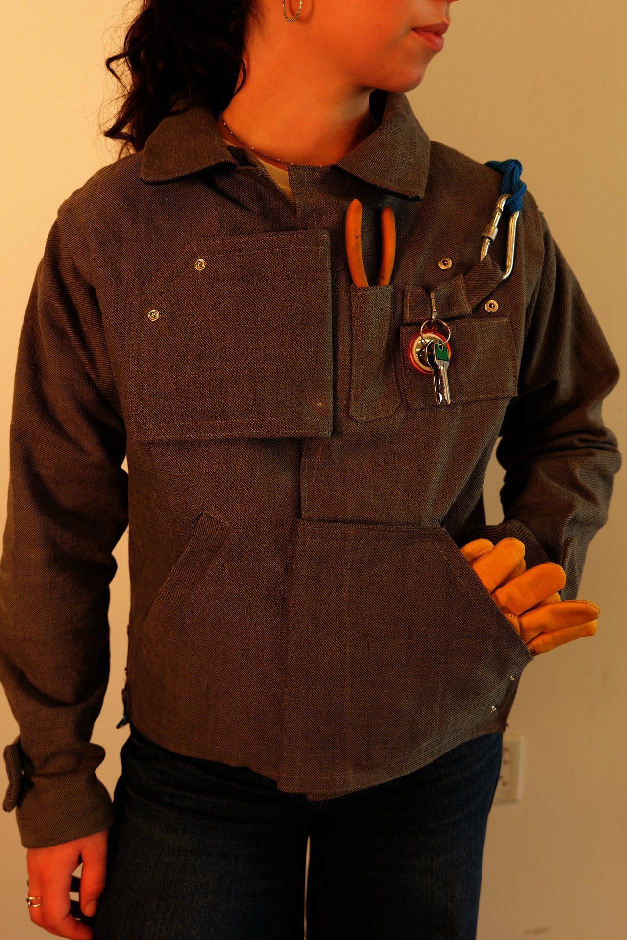 The Open Loop Jacket. A girl wearing a gray, long-sleeve carpenter's jacket with many pockets that hold utilitarian tools. The sleeves of the jacket snap off, and pockets act as the upper and lower closures.