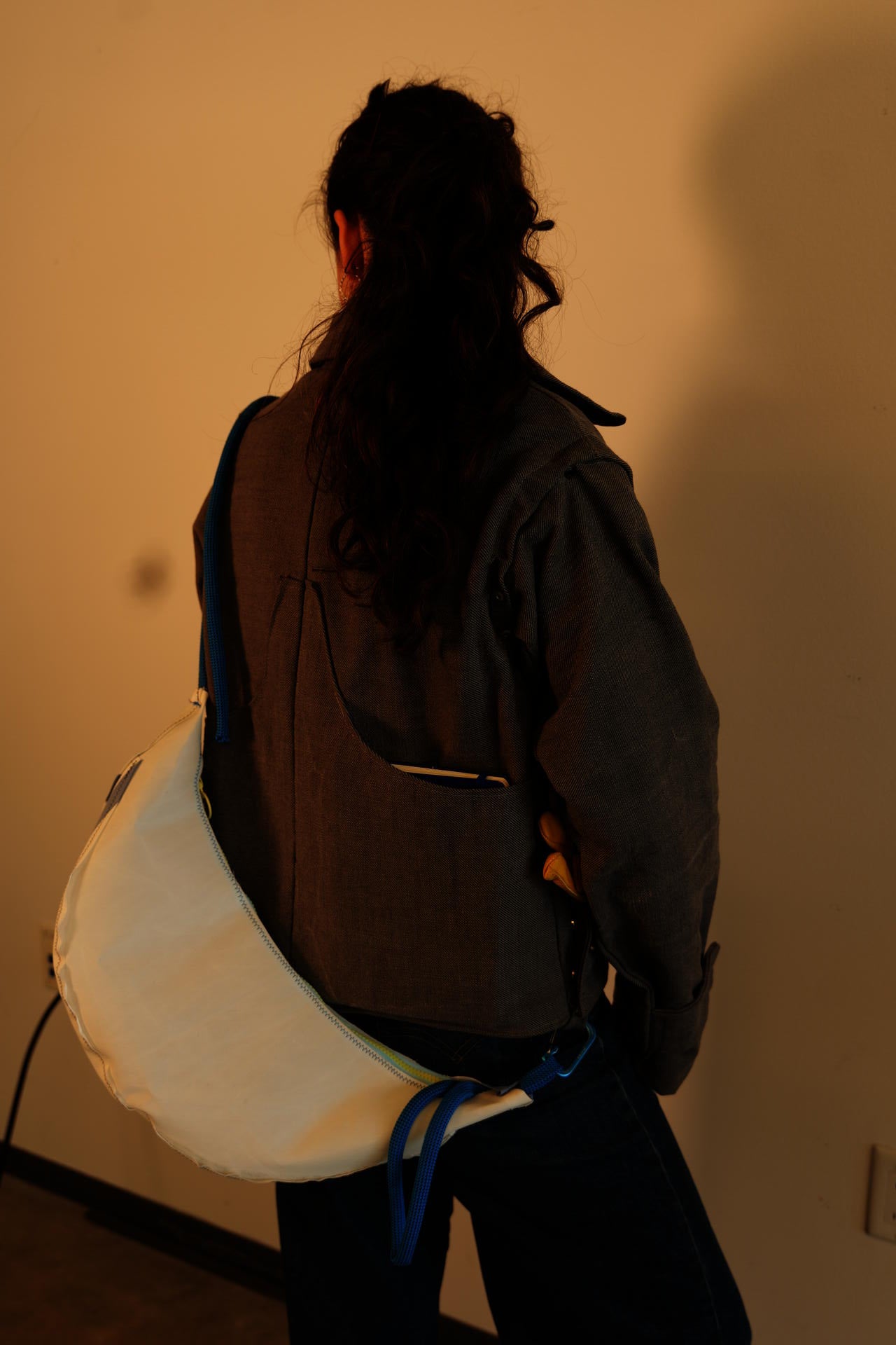 The back of a girl wearing a gray jacket and blue jeans. A white crescent bag with blue rope straps has been hooked into loops on the jacket to sling it across the back. A notebook is placed in the right side panel.