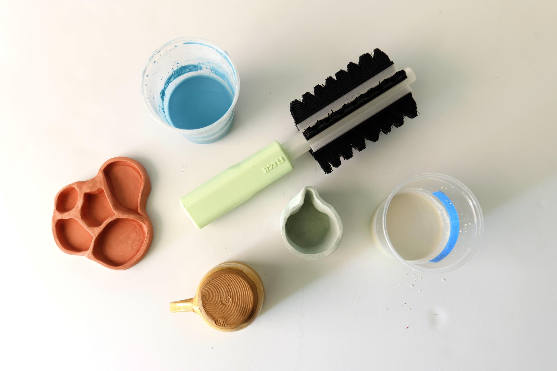 On a white background, a green handled brush with black bristles oriented near several jars of blue, grey, and white ceramic glaze liquid. A yellow mug.
