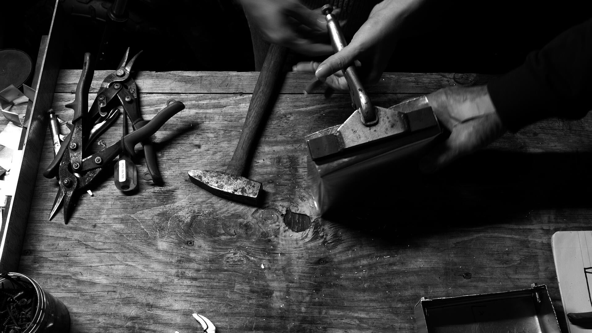 Video still in black and white from a project about the passing of generational knowledge and skill. Two people are shaping a metal box with sheet metal tools.
