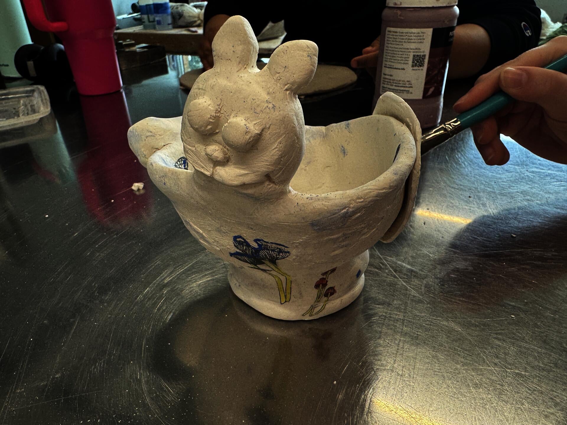 A student out of frame glazes an oversized tea cup with mushrooms and a cat face