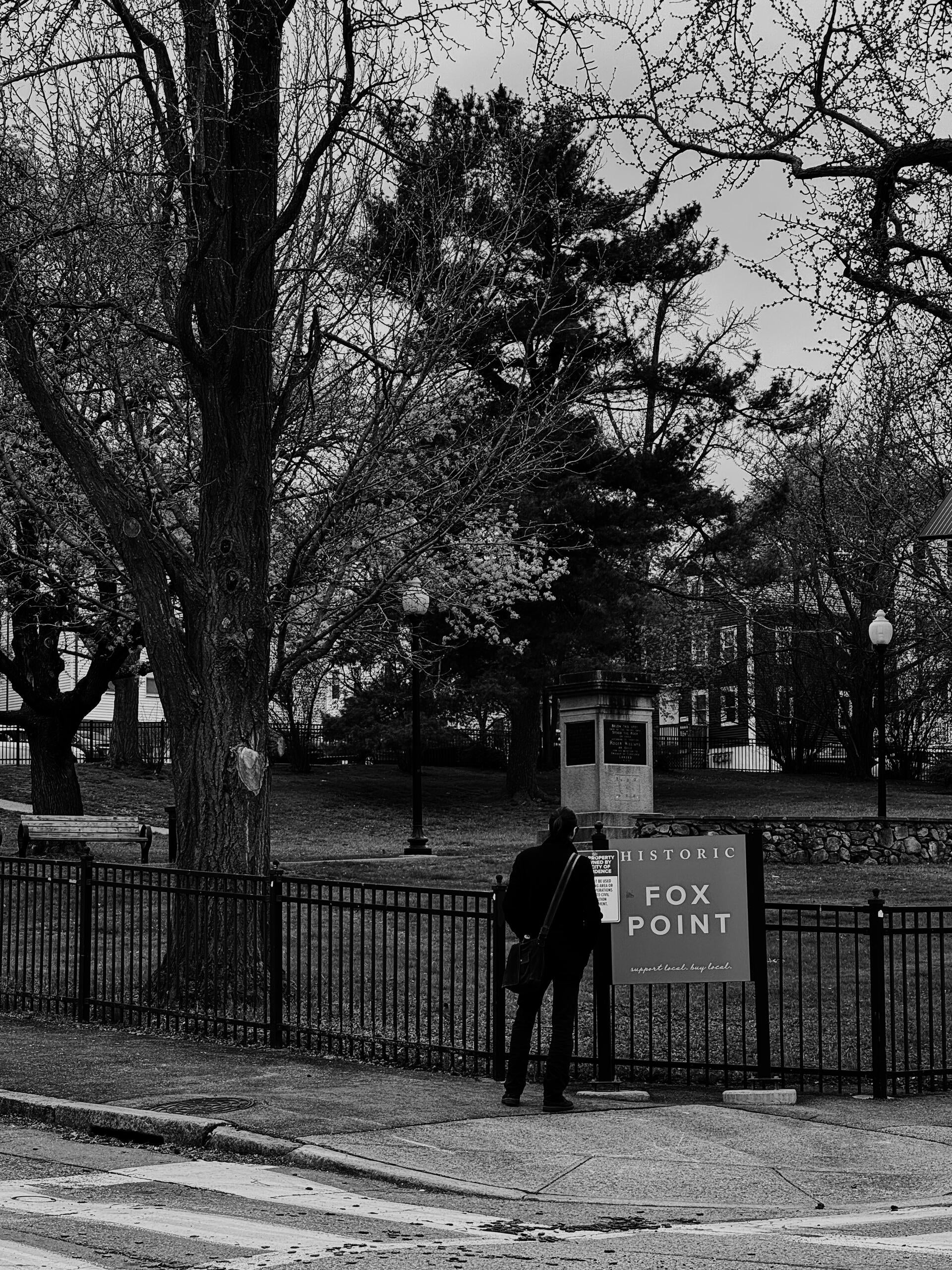 A black-white photo with a man standing back to the camera, looking at a billboard describing an urban green space in the background. He is wearing a black jacket with his hair tied up.