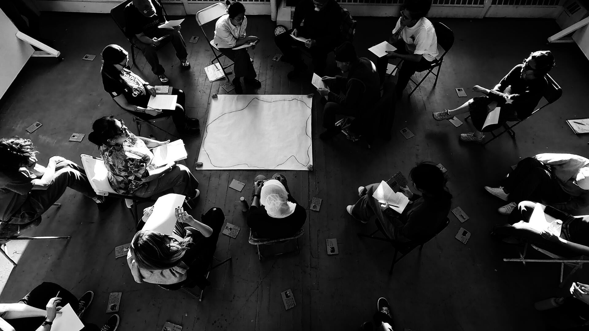 A high-angle black-white shot with 18 people sitting in a circle listening and reading. Three of them are holding printed paper and reading text from it. Paper and cardboards with text and images are scattered around.
