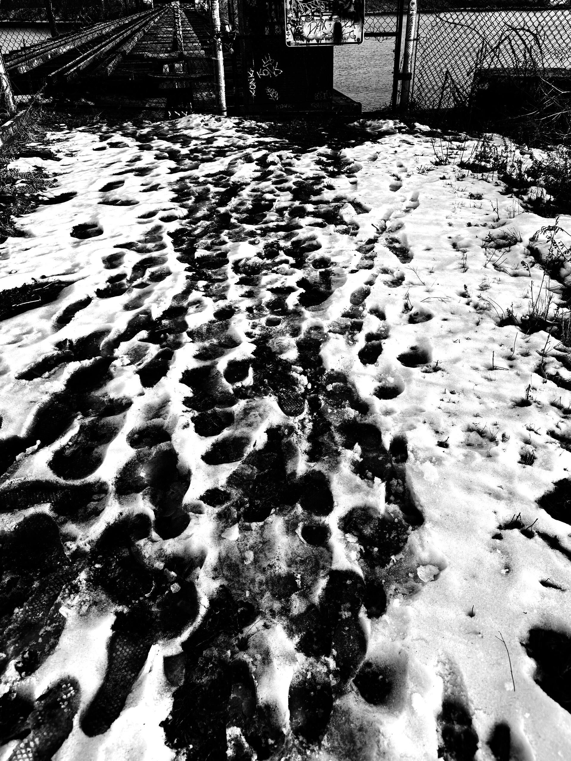 Black-white photo with footprints on the snow pathway directing to a half-open broken iron gate in the distance. Inside the gate, train tracks on a bridge are forwarding.