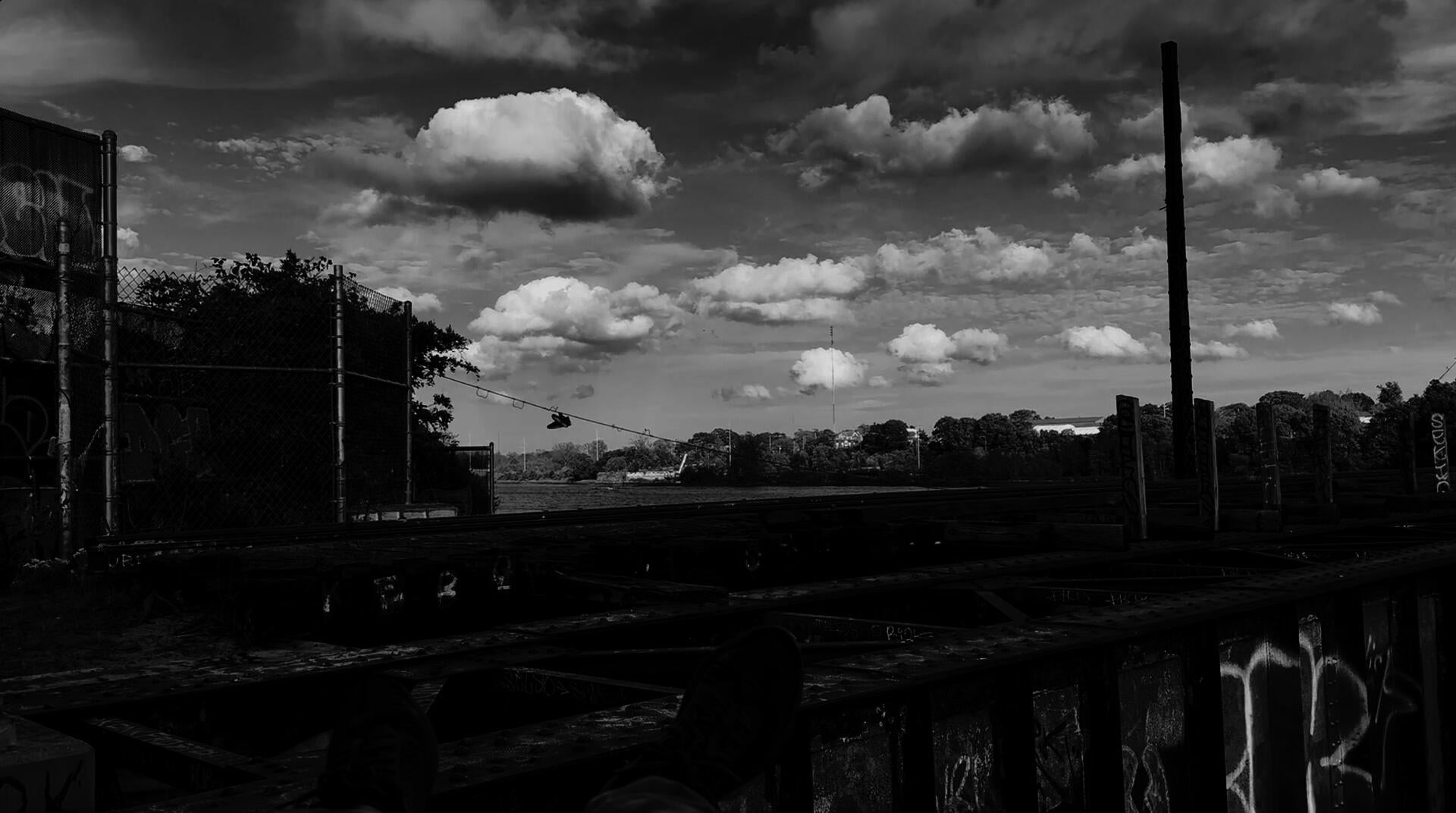 Black-white photo taken from a person’s angle watching over the train track running through the middle of the frame, with feet waving in the foreground. In the distance, a pair of shoes hangs from a rope.