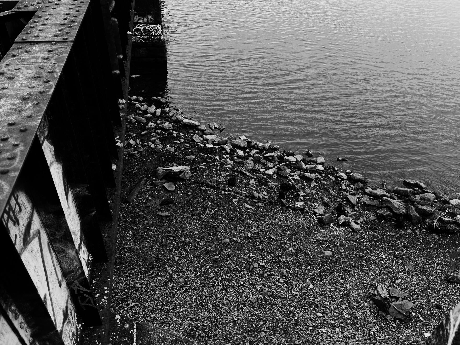 An overhead black-white shot of a rocky shore on the far side of the frame while a part of the train track is leading towards on left front side.