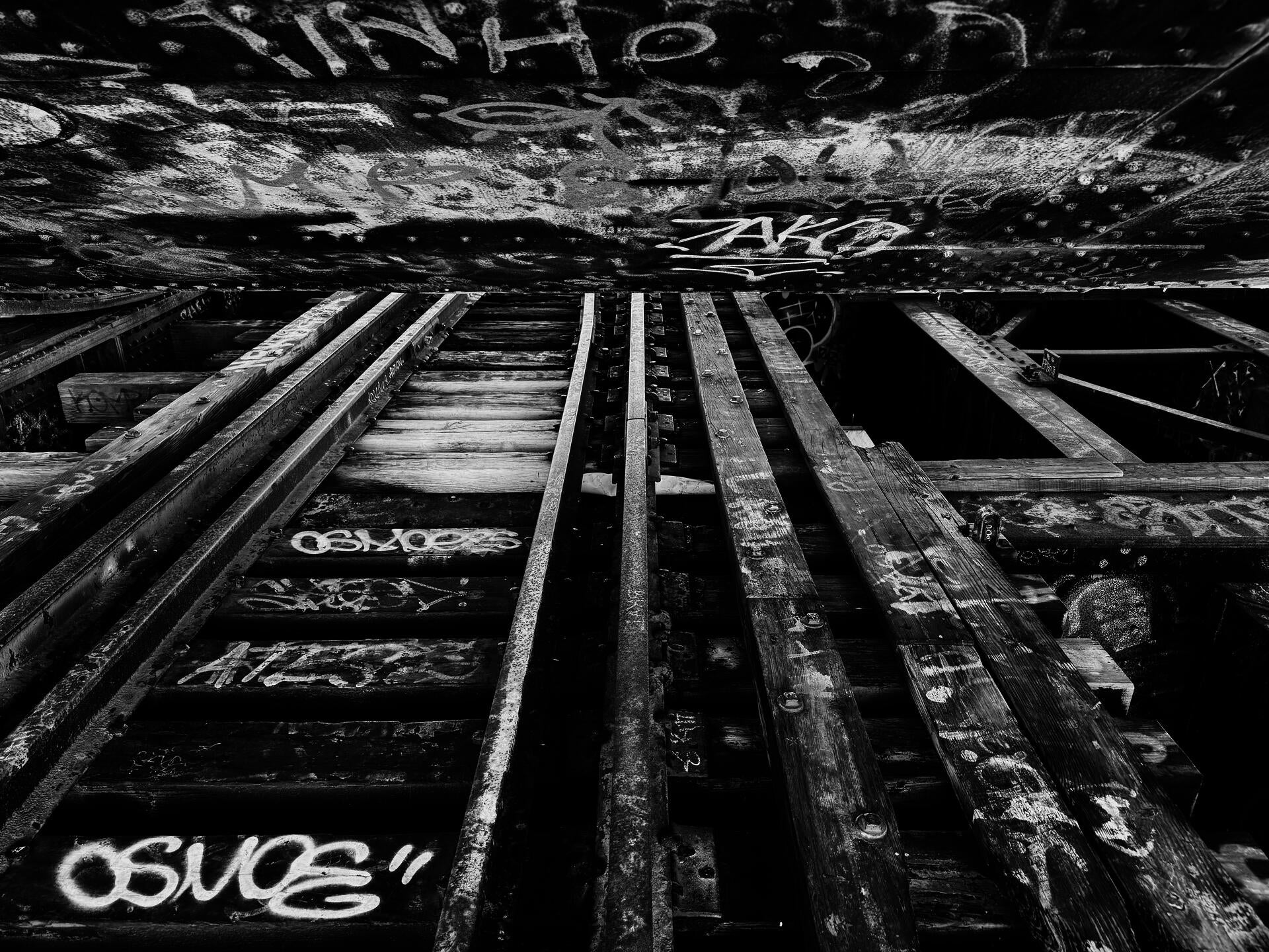 Black-white photo with a stretch of old, abandoned train tracks under an up-structure of a bascule bridge, covered with graffiti and burnt woods. A white sheet of paper is rolled up and embedded in the middle.
