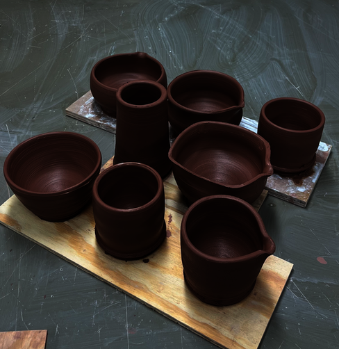 assorted thrown greenware pottery in dark brown clay, resting on dusty table.