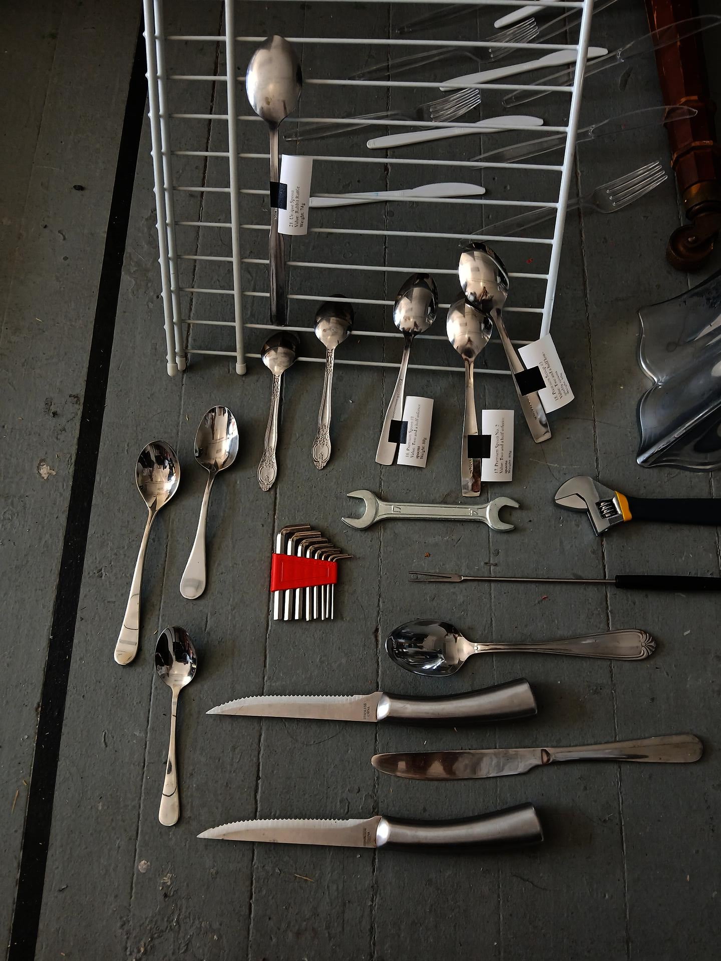 Assorted spoons, knives, and utensils arranged on a gray tiled floor beside a white drying rack, with plastic cutlery, hex keys, a wrench, and an adjustable spanner, some with white labels.