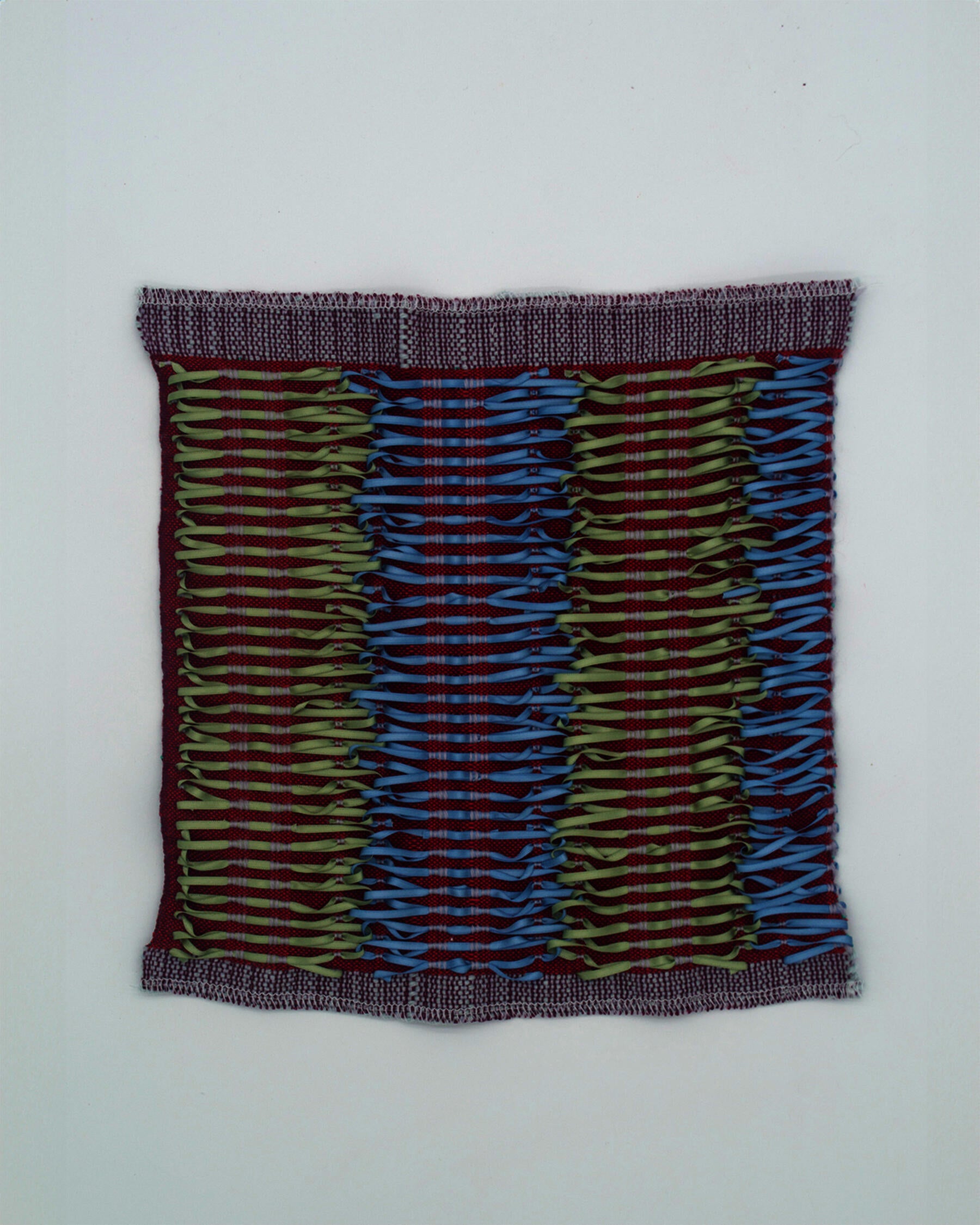 A maroon woven fabric with warm green and baby blue ribbons tied onto the surface. The ribbons run vertically, interlocking in a repeating pattern reminiscent of corset lacing