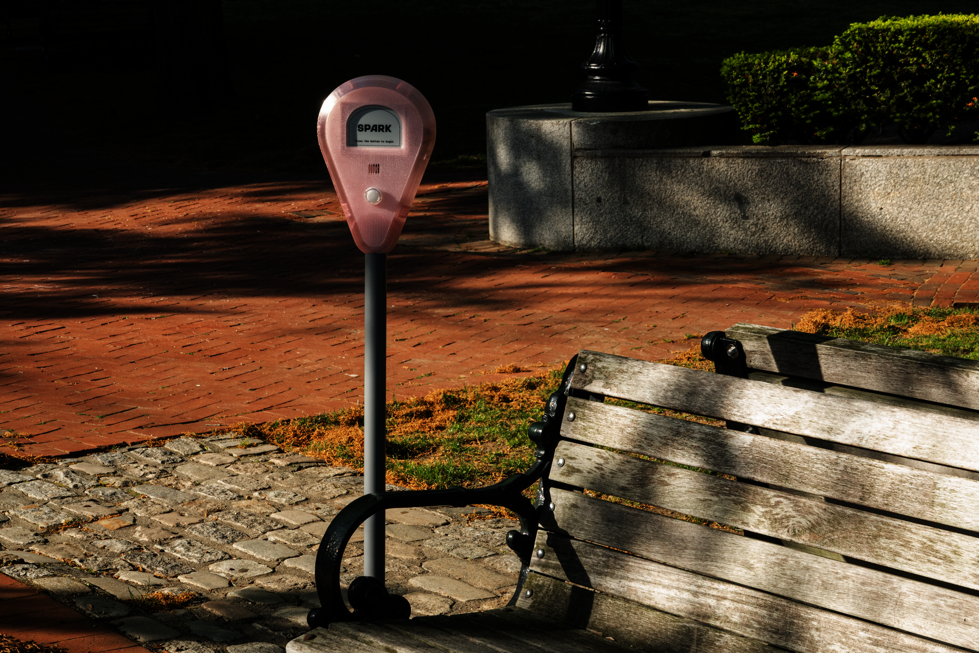 Spark device installed beside a park bench on a brick walkway, with trees, grass, and sunlight in the background.