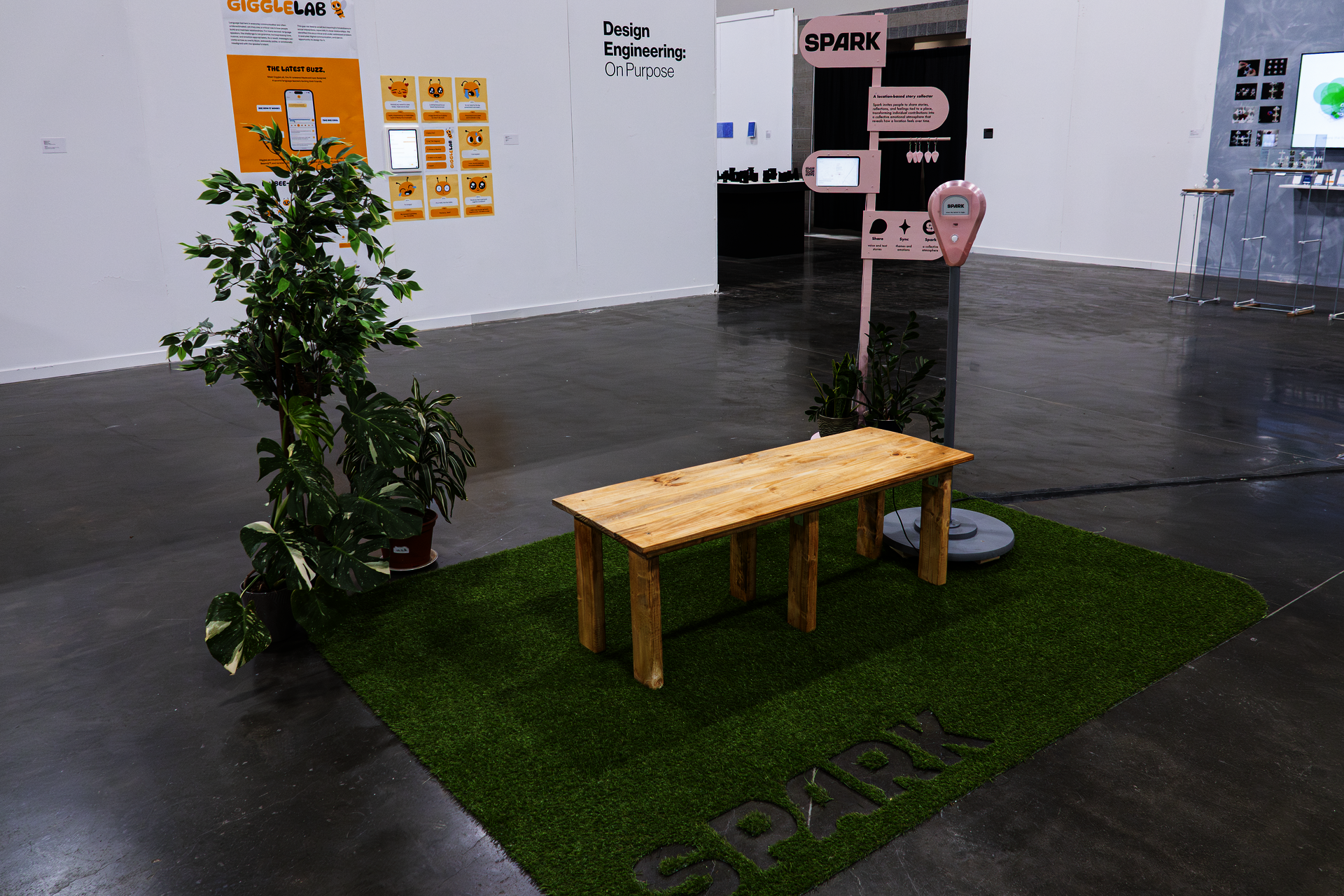 Angled view of Spark exhibition setup with artificial grass, wooden bench, plants, and signage.
