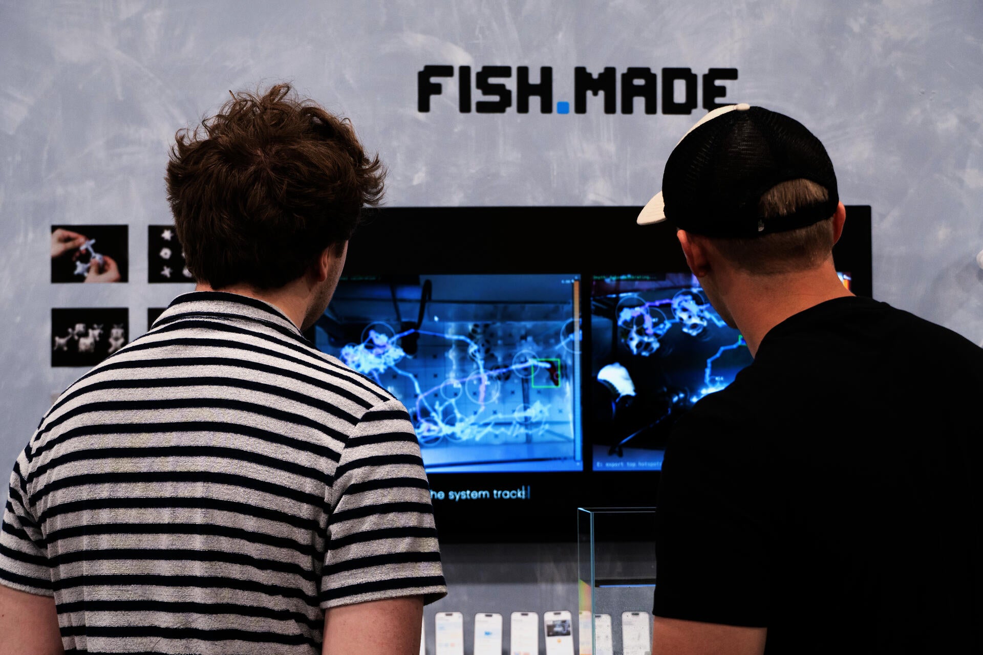 Two visitors view the FISH.MADE installation, which shows fish movement tracking visuals on a screen alongside the project logo and physical prototype display.