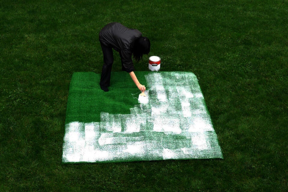 Photograph of a woman on grass painting astro turf with white house paint