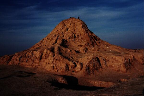 Color photograph of the Assur Ziggurat. What is left of the ziggurat (it is mostly eroded) rises out of sand in a big mound against a blue sky. There are three small figures standing on top.
