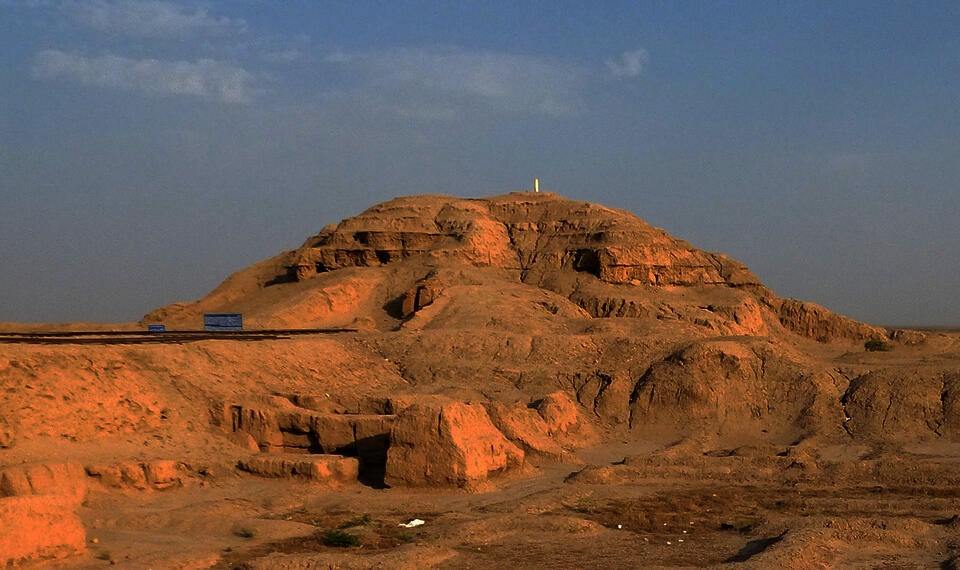 color photograph of the Anu Ziggurat at Uruk. The remains of the stepped pyramid ziggurat emerge from the yellow sand desert, with blue sky above.