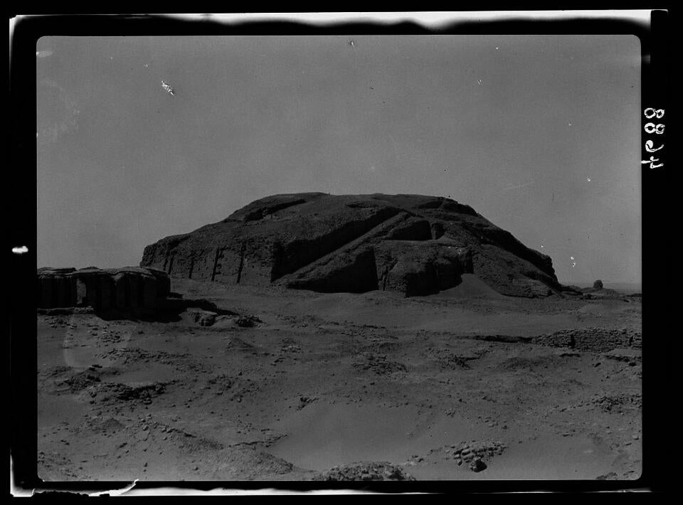 Black and white photograph of the Ziggurat of Ur. The remnants of the ziggurat sit on the horizon line, with sand below and sky above. Only the ziggurat's square base remains, with a triangular staircase.