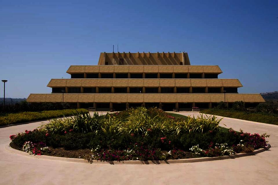 color photograph of the Chet Holifield Federal building in California. Taken from a low angle, there is a lush green planting in the forefront of the composition. In the background, the building, a modernist interpretation of a ziggurat, rises from the horizon line against a blue sky.