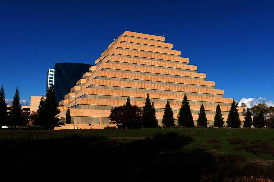 Color photograph of the California Department of General Services building, a 10-story contemporary ziggurat building. Taken from a 3/4 view, this photograph shows of the stepped sides of the tan building against a blue sky.