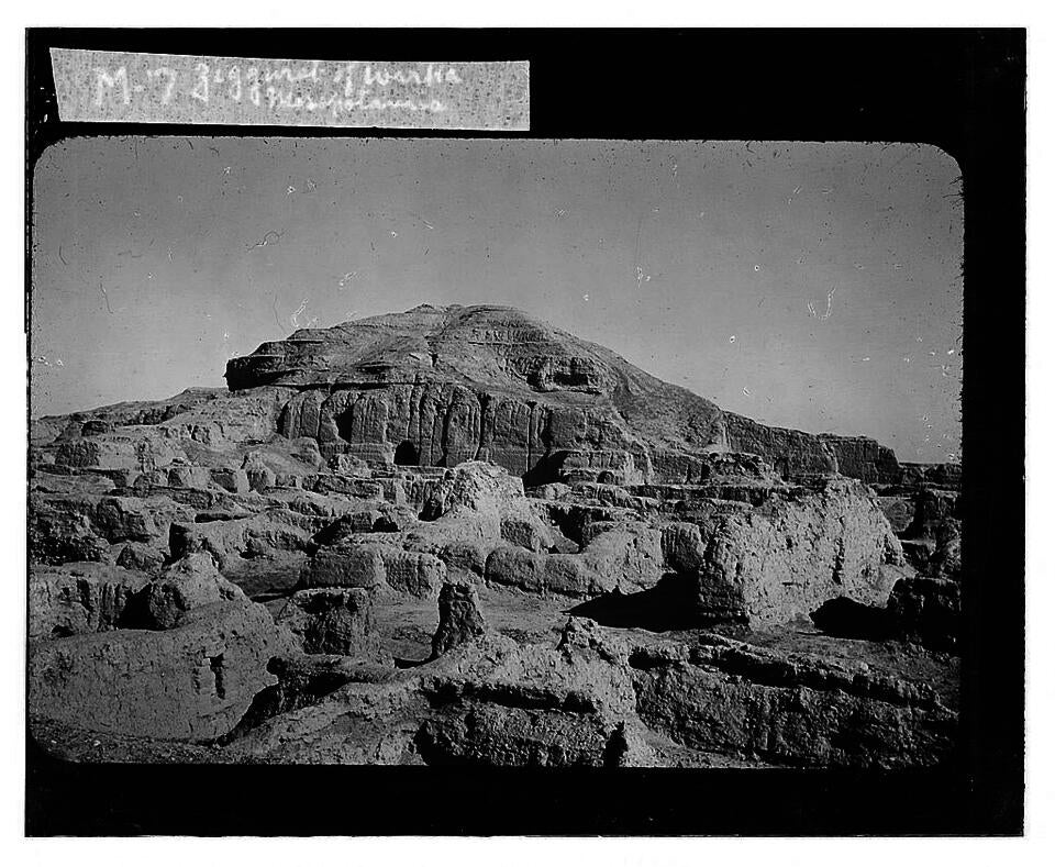black and white photograph of the Ziggurat of Warka. Foreground is scattered stone blocks in a desert landscape. Beyond is the ruins of the Warka Ziggurat.
