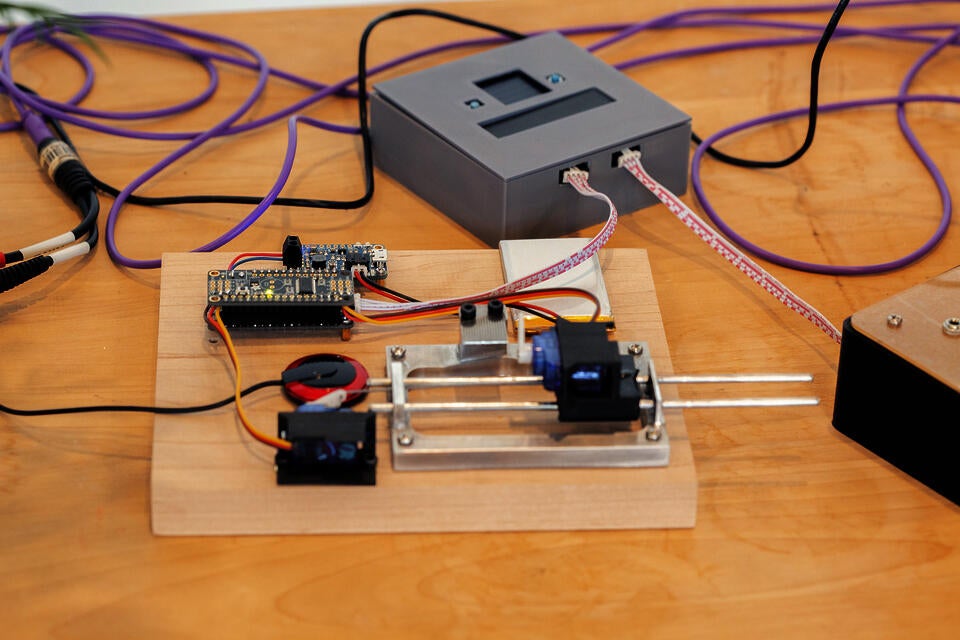 A close view of a robotic music box. A small motorized plastic lever is angled upward facing the music box tines. In the background, a small gray box can be seen. Wires connect the box and a circuitboard on the robotic music box.
