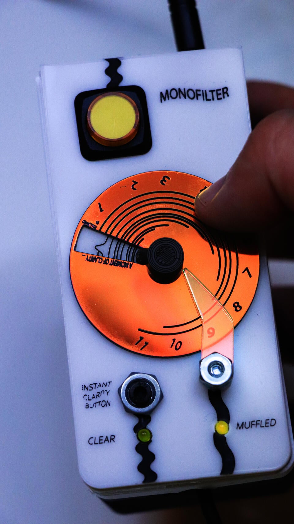 photograph showing a hand holding the Monofilter (roughly the size of a remote, but deeper). The top left corner has a large toggle-button (on/off), the center has a large flat circle that rotates: a dial to change the "muffling duration" (an time interval between each moment of musical clarity and the next). below the dial is an "instant clarity" button, and two lights to indicate whether the sound is "clear" or "muffled". A thumb is placed on the dial.