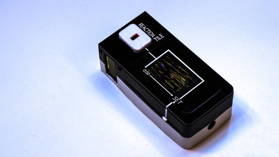 Photograph of the reaction tile: a device about the size of a remote but thicker (1.5inches total). The top surface has a heart rate sensor located near the top (for the thumb). The bottom 2/3 of the top surface is occupied by a flap (like casette players) to place the tile in. The side has a small screen and a slider control to set the total duration to sync the engraving speed/ song duration.