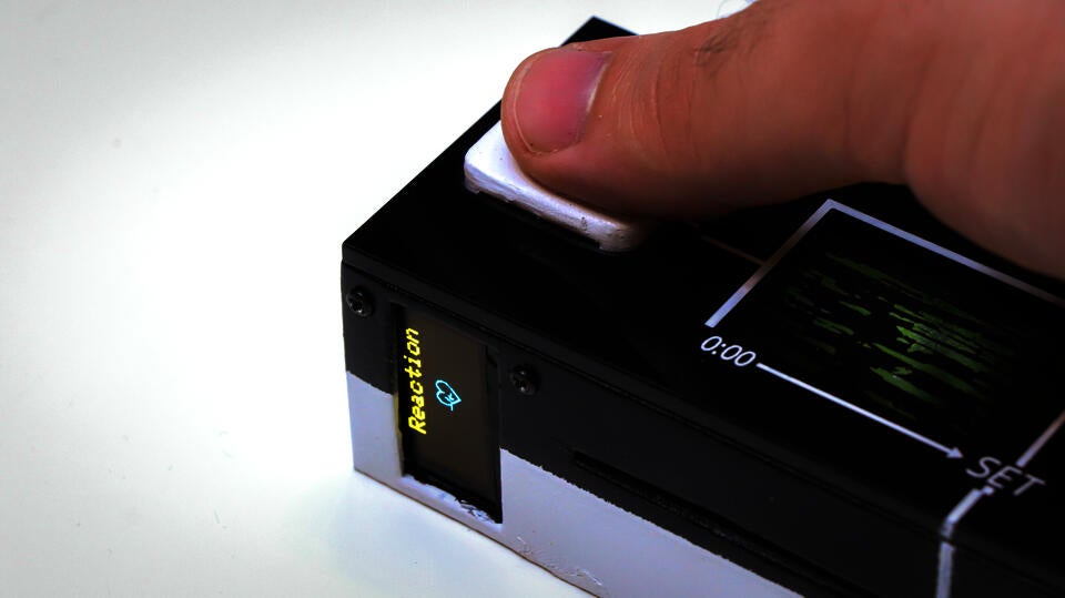 Closeup photograph showing the Reaction Tile (device, roughly the dimensions of a remote but about an inch and half thick) held by a hand. The thumb is on a heart rate sensor (top surface), below is where the tile is placed for engraving: the device opens up like a cassette player. A screen on the side has a small screen showing "reaction" and a cartoon heart.