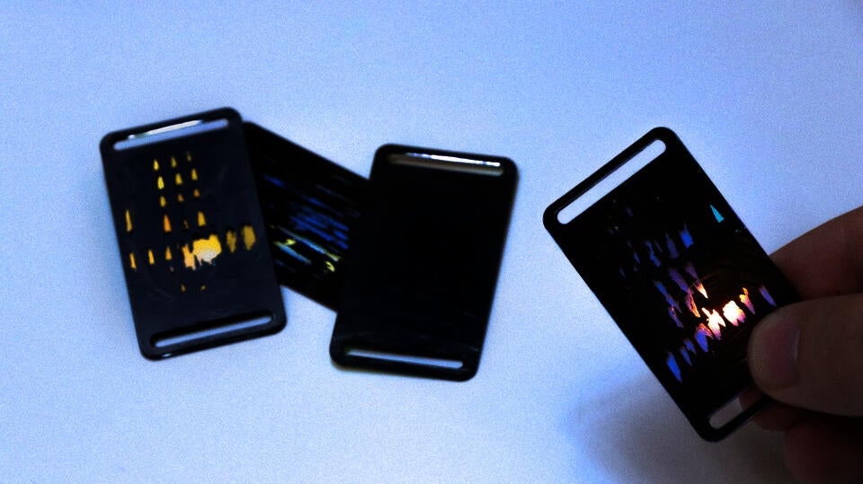Photograph showing three (domino-sized but 1/8 inches thick) "tiles", each is black (thin layer of paint) but with visible light reflections underneath, as a result of engraving that scratched off some paint in reaction to heart beat variability. On the right side of the image is a hand holding up a tile. bordering the thinner edge of each tile is a thin (0.1in) slot that stretches along; this provides ways to secure tiles to other things (eg a frame, tied to a string, shoelaces?)