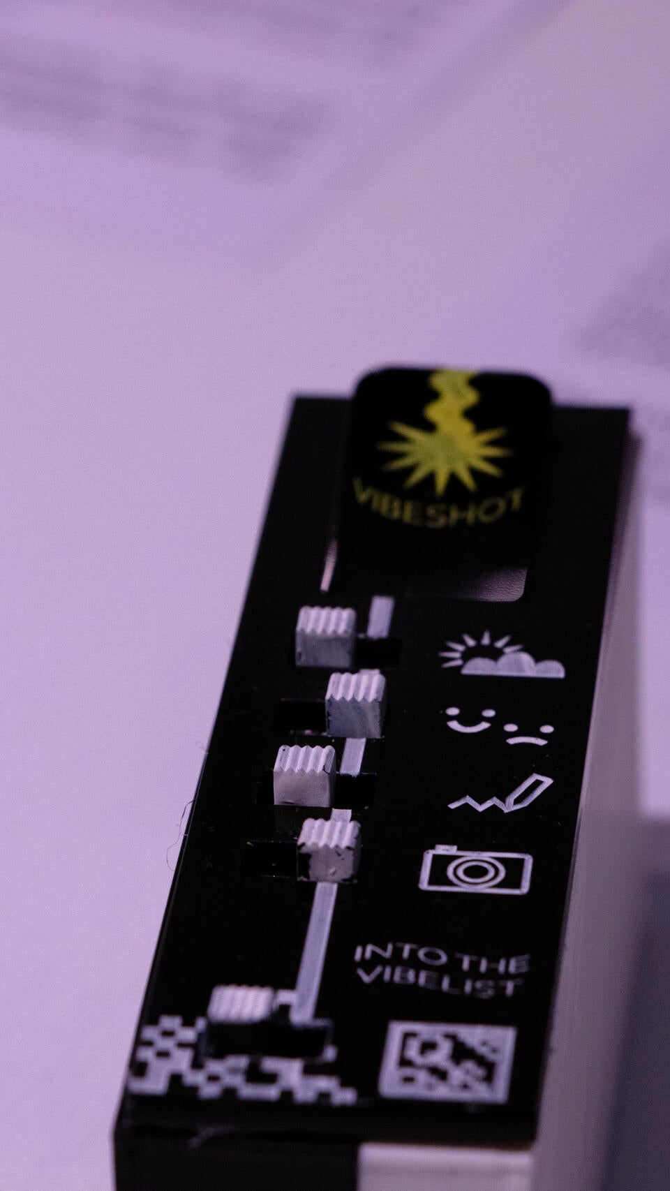 Photograph showing the Vibeshot feature: which is on the side surface of the device. The top of this face has a button labeled "vibeshot". From the button stretches a line which is interrupted by five toggle switches; symbolizing whether the elements the toggles represent are capture in the vibelist. the elements (top to bottom) are weather+location, self-reported feeling, text, image and a web-link (to share with others)