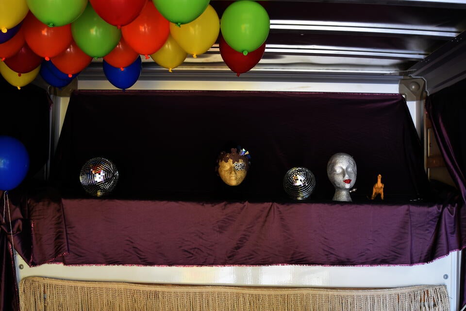 Interior of a U-haul, draped in shiny purple fabric, with two disco balls and two foam mannequin heads and a toy dinosaur sitting on a ledge. Balloons hover on the ceiling.