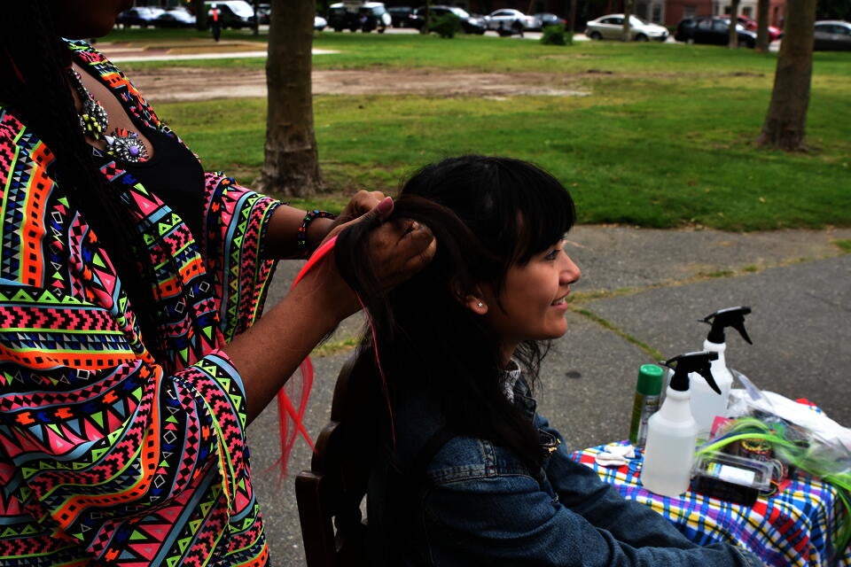 Female teen sits as another female teen in a bright shirt and necklace braids a bright pink hair extension into her hair. They are outside in a park.
