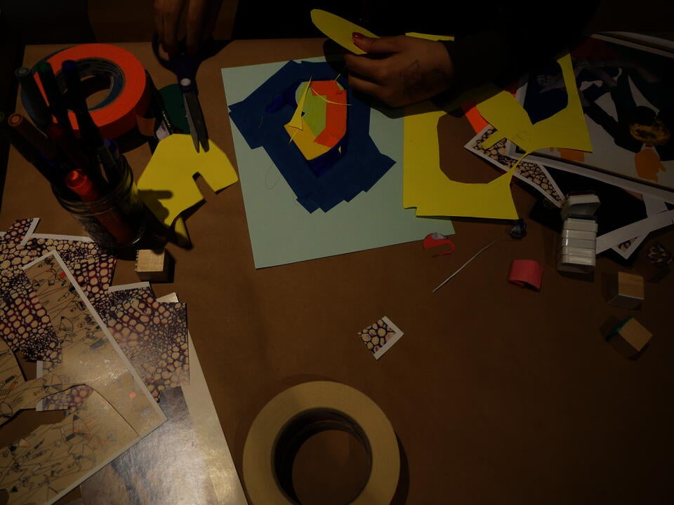 overhead color photo of a table covered in brown paper, strewn with colorful art supplies; a pair of hands grasp a yellow paper circle and a pair of scissors in the top of the image