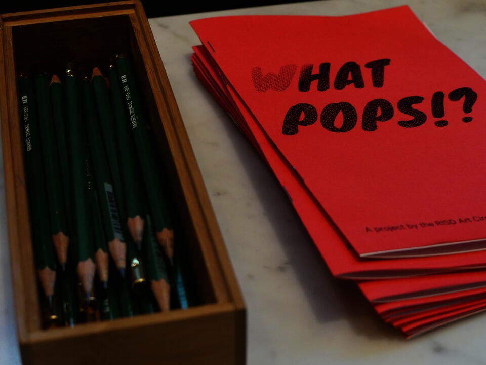 close-up color photograph of a box of green pencils and pile of salmon colored booklets that say 'What Pops!?'