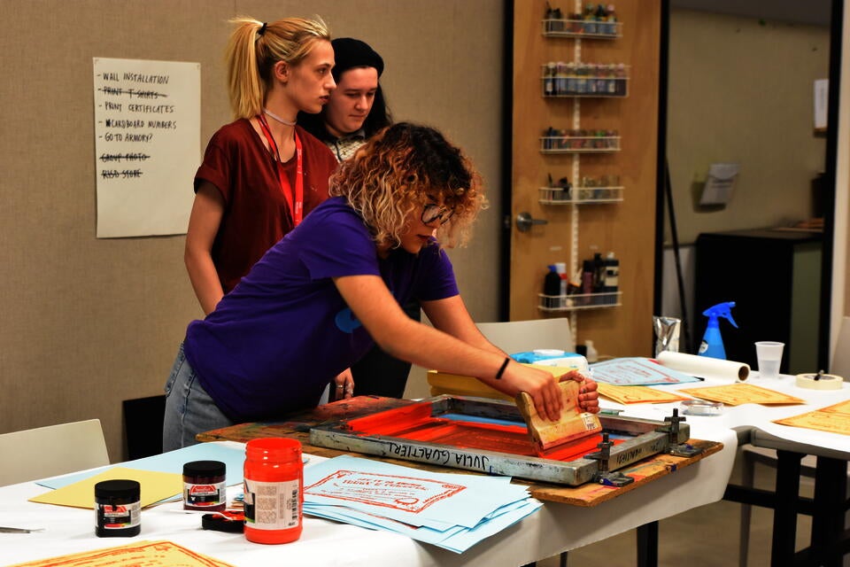 Three teens screenprinting paper with bright orange ink.