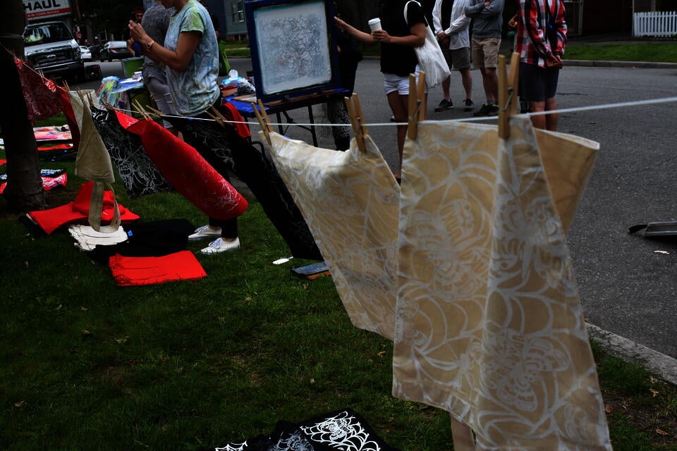 a clothesline of canvas totebags and red and black handkerchiefs hand to dry after being screen printed on. People stand in the background, near a screen printing screen. 