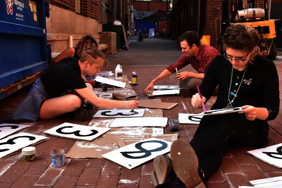 Three teens and one adult male sit on the ground in an alleyway, painting large black numbers onto pieces of cardboard.