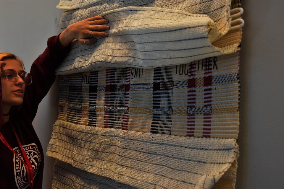 A young woman in a RISD sweatshirt lifts the fold of a hanging woven textile with several flaps
