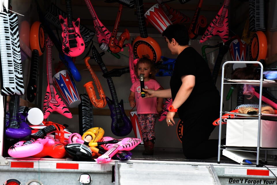 An adult holds a microphone up to a small child in a pink shirt and cowboy boots. They are in the back of a U-haul that is filled with colorful inflatable rock instruments.