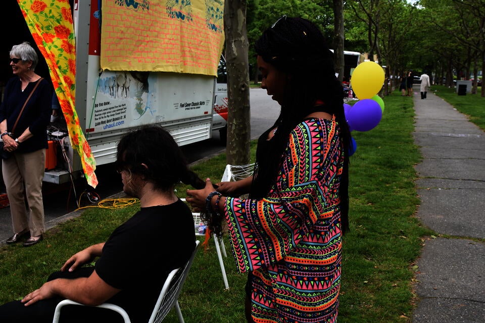 A black teen with long braids and a colorful shirt braids the hair of a seated teen. They are outside in a park near a U-haul with a large mural on it.