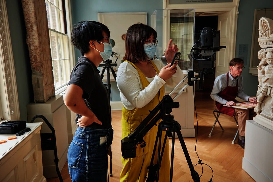 Two masked young people film in a museum gallery. They look at a carved stone work, tripods standing beside and behind them. Another person is seated nearby. 