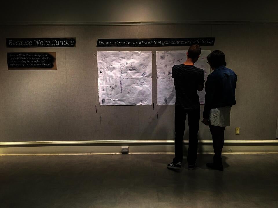 Two figures stand and look at drawings on the sheets of paper hung on the wall, under the sign 'Draw or describe an artwork that you connect with today.'