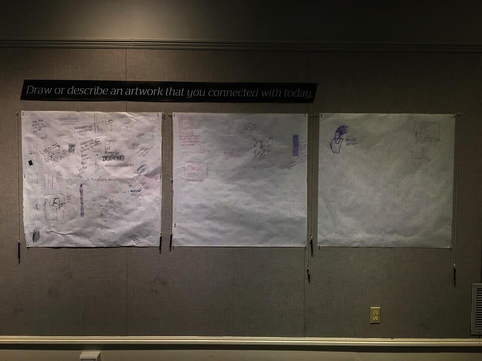 Three sheets of paper covered in drawings hang beneath a sign that says 'Draw or describe an artwork that you connect with today.'