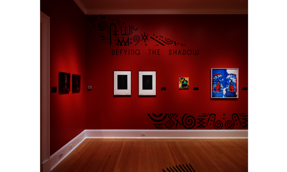 Gallery with red walls hung with several works. The wall is embellished with black designs and text that reads: “Defying the Shadow.”