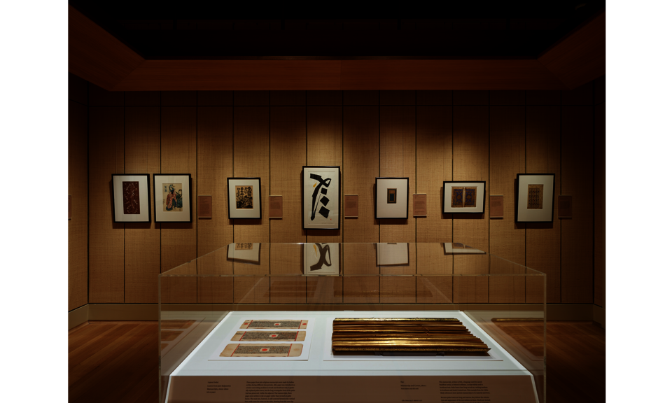 Gallery space with tan walls and peaceful lighting. Several works hang on the far wall, with additional objects in a case in the foreground.