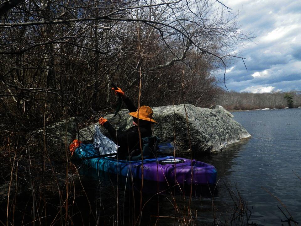 A person seated in a kayak on calm water. The trees above them and the vegetation we view them through are leafless. The sky is cloudy.