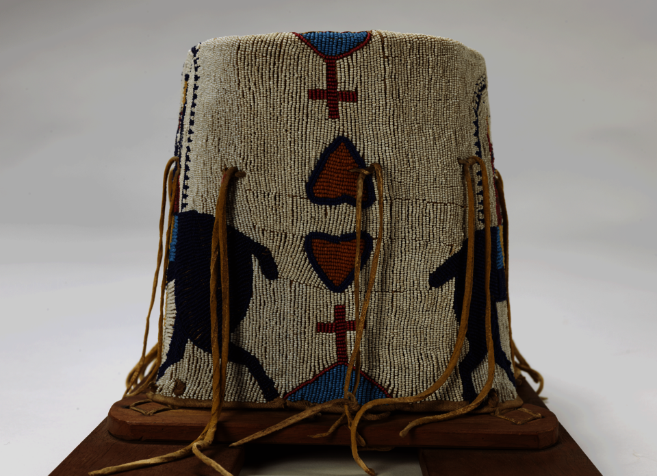 Detail of beaded object with leather ties hanging down and wood supports at bottom. White background beaded with pink and blue hearts and red and blue crosses.  