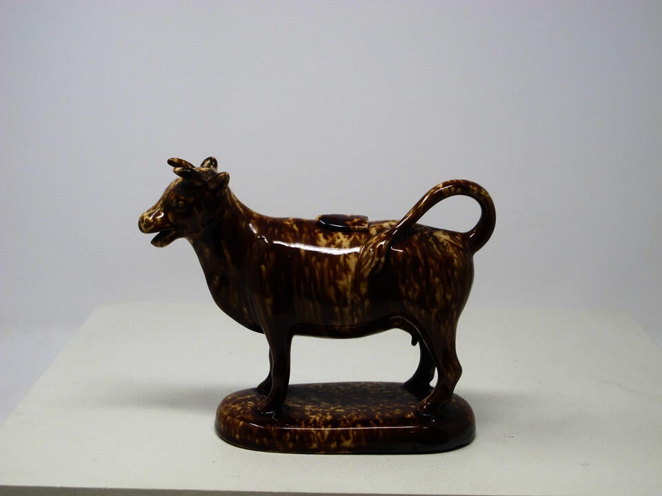 A brown sculptural creamer in the shape of a cow with horns. There is a small flat lid in the center of its back.