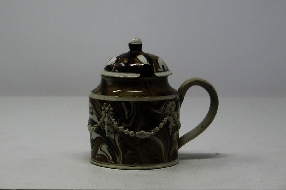 An earthenware mustard pot that is white, light brown and dark brown. The lid and handle are rounded and there are swag floral decorations on the body.
